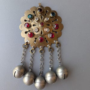 May include: A circular brass brooch featuring a scalloped edge and a central floral pattern. The piece is adorned with inset red, green, and clear faceted stones. Five silver-toned chains hang from the bottom, each ending in a round metal bell.