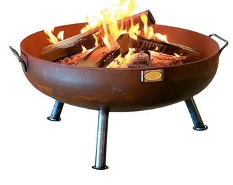 May include: A round, rust-colored metal fire pit bowl with three sturdy legs and two side handles. The bowl contains burning logs with bright orange and yellow flames. A small, rectangular metallic plaque is attached to the front rim.