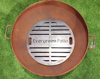 Puede incluir: Un pozo de fuego de metal oxidado con un inserto de rejilla redonda de metal. La rejilla tiene una serie de ranuras paralelas y el texto "Evergreen Patio" es visible en la rejilla.