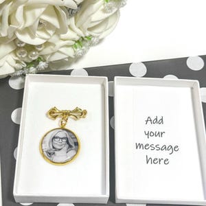 May include: A gold brooch with a circular pendant displaying a black and white photograph of a person wearing spectacles. The brooch is presented in a white box, beside another box with the text "Add your message here". A bouquet of white roses is in the background.