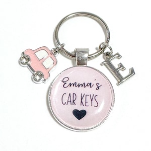 May include: A silver keyring with a pink car charm, a silver letter "E" charm, and a round charm that says "Emma's CAR KEYS" with a black heart. The round charm has a pink background.