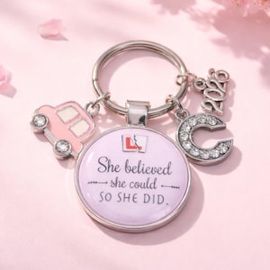 May include: A silver keyring with a round, light purple charm that reads "She believed she could so she did." The keyring has a pink car charm, a rhinestone-studded letter "C", and a "2022" charm. Pink flowers and petals are in the background.