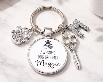 May include: A silver keychain with multiple charms. The charms include a paw print, scissors, and the letter "M". The main charm has the text "AWESOME DOG GROOMER Maggie". The paw print and letter "M" are embellished with small, clear crystals.
