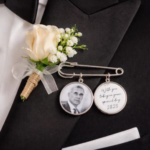 May include: A cream rose and white flower boutonniere pinned to a black tuxedo jacket. A silver pin holds two circular charms, one with a photo and the other with the text "With you today on your special day 2823."