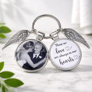 May include: A silver keyring with two circular pendants. One pendant displays a black and white photograph of a couple, the other has the text "Those we love are always in our hearts." Wing charms are attached.