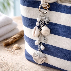 May include: A beach-themed keyring featuring seashell, pearl, and letter "F" charms. The keyring is attached to a blue and white striped tote bag, creating a coastal theme. The charms are silver-toned and the bag has a nautical design.