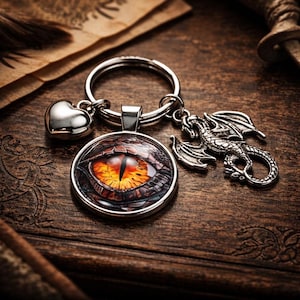May include: A silver-toned keyring featuring a circular pendant with a detailed orange and black dragon eye graphic under a glass dome. The keyring includes a small polished silver heart charm and a detailed silver dragon figure charm attached.