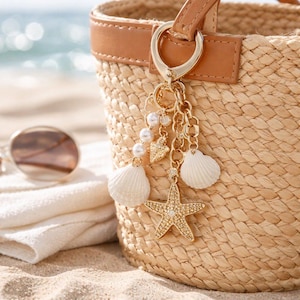May include: A woven straw tote bag with a brown leather handle and a gold-coloured charm. The charm features a starfish, seashells, pearls, and heart-shaped pendants. The bag is on a sandy beach.