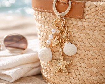 May include: A woven straw tote bag with a brown leather handle and a gold-colored charm. The charm features a starfish, seashells, pearls, and heart-shaped pendants. The bag is on a sandy beach.