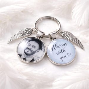May include: Silver keyring with two circular pendants and two wing charms. One pendant shows a black and white photo of a smiling person. The other pendant has the text "Always with you" with a heart.