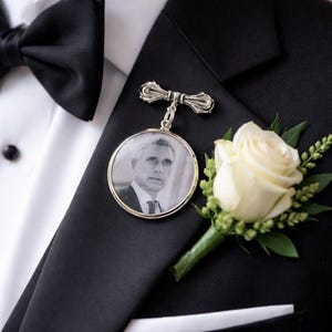 May include: A black tuxedo with a white dress shirt, a black bow tie, and a silver memorial pin with a photo. A white rose boutonniere with green leaves is also visible. The image is a close-up of formal attire.