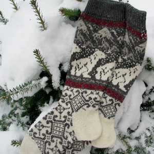 May include: A pair of white and gray knitted socks with a red and white geometric pattern. The socks are folded and placed on a snowy evergreen tree branch.