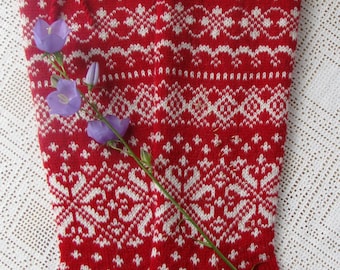 May include: A pair of red and white knitted socks with a traditional Scandinavian pattern. The socks are folded over at the top and have a white flower stem draped over them.