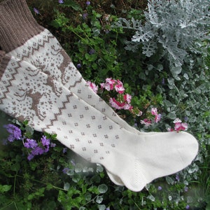 May include: A pair of white knitted socks with a brown geometric pattern. The socks are lying on a bed of green plants and flowers.
