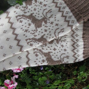 May include: A pair of knitted socks with a white background and brown geometric pattern. The socks are folded and lying on a bed of green plants and purple flowers.