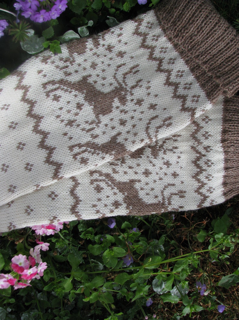 May include: A pair of knitted socks with a white background and brown geometric pattern. The socks are folded and lying on a bed of green plants and purple flowers.