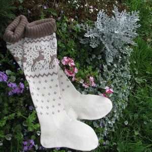 May include: A pair of white knitted socks with a brown reindeer pattern. The socks are lying on a bed of green grass and pink flowers.