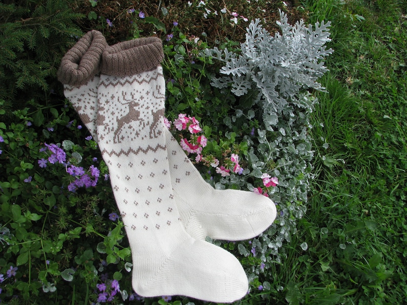 May include: A pair of white knitted socks with a brown reindeer pattern. The socks are lying on a bed of green grass and pink flowers.