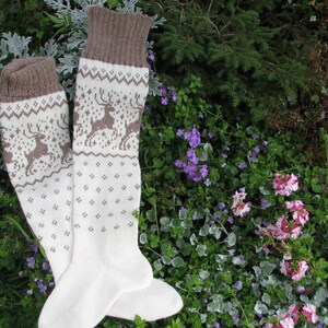 May include: A pair of white knitted socks with a brown reindeer pattern. The socks are folded and lying on a bed of green and purple flowers.