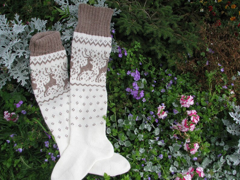 May include: A pair of white knitted socks with a brown reindeer pattern. The socks are folded and lying on a bed of green and purple flowers.