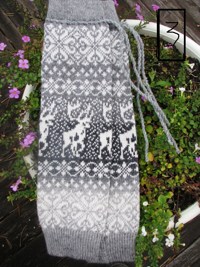 May include: Grey and white knitted leg warmers with a geometric pattern and a white background. The leg warmers have a fringe detail at the top.