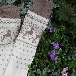 May include: A pair of white knitted socks with a brown reindeer pattern. The socks have a brown cuff and are lying on a bed of green and purple flowers.