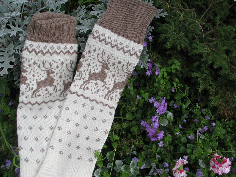 May include: A pair of white knitted socks with a brown reindeer pattern. The socks have a brown cuff and are lying on a bed of green and purple flowers.