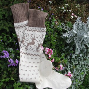 May include: A pair of white and brown knitted socks with a reindeer pattern. The socks are lying on a bed of green and purple flowers.