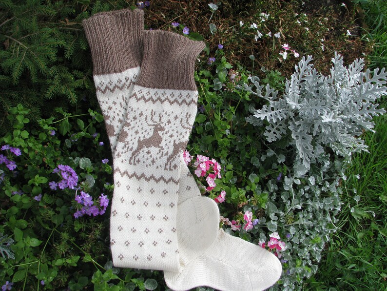 May include: A pair of white and brown knitted socks with a reindeer pattern. The socks are lying on a bed of green and purple flowers.