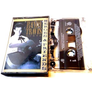 Puede incluir: Una caja de cassette negra con una ventana de plástico transparente que muestra la cinta en el interior. La parte frontal de la caja presenta una foto de Randy Travis, un cantante de música country, sosteniendo una guitarra. El texto en la caja dice "Randy Travis Heroes & Friends Duets".