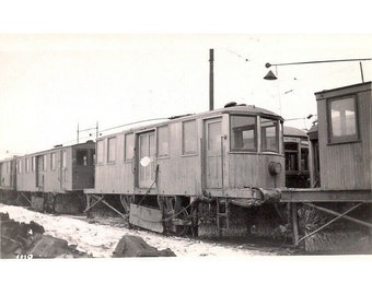 Peut inclure: Une photo en noir et blanc d'un train à neige vintage. Le train est en bois et possède une grande lame à l'avant. Le train est sur une voie enneigée et il y a d'autres wagons derrière lui.
