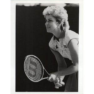 Puede incluir: Una foto en blanco y negro de una mujer jugando al tenis. Lleva una camisa blanca con un patrón a rayas y una falda corta. Sostiene una raqueta de tenis en su mano derecha y está mirando hacia arriba a la pelota.