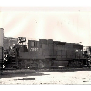 May include: A black and white photo of a large, gray diesel locomotive with the number 7964 on the side. The locomotive is on a set of railroad tracks. The locomotive has a large, black exhaust stack and a large, black cab.