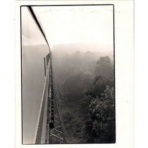 Pode incluir: Uma fotografia a preto e branco de uma ponte ferroviária. A linha férrea está do lado esquerdo da imagem e a ponte é feita de vigas de metal. A ponte está sobre uma floresta. A floresta está coberta de nevoeiro.