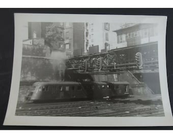 May include: A black and white photograph of a train traveling through a city. The train is passing under a bridge and there are buildings in the background.