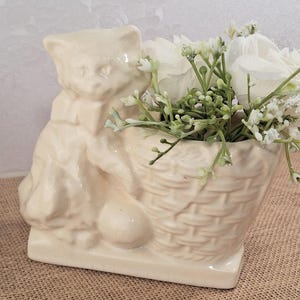 May include: A cream-colored ceramic cat figurine holding a woven basket filled with white flowers and greenery. The cat is seated, with a bow around its neck, and a ball at its feet. The figurine is on a rectangular base.