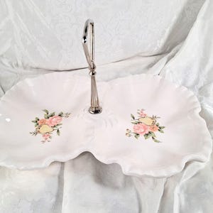 May include: White ceramic serving tray with a silver handle. The tray has a two-tiered design with a wavy edge and is decorated with pink roses and green leaves. A decorative piece for serving food or displaying items.