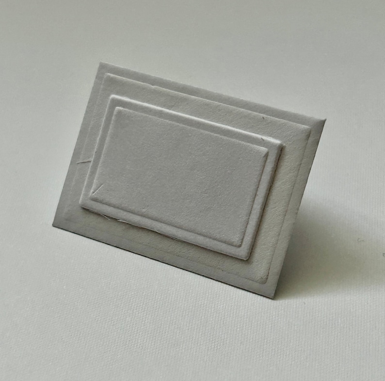 Puede incluir: Una tarjeta blanca con un marco rectangular en relieve. El marco tiene tres capas, creando un efecto de profundidad sutil.