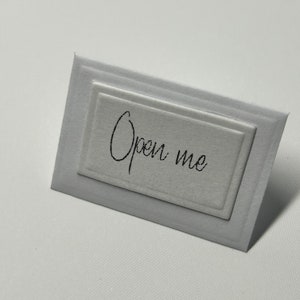 Puede incluir: Una tarjeta blanca con un borde en relieve y el texto "Open me" escrito en negro.