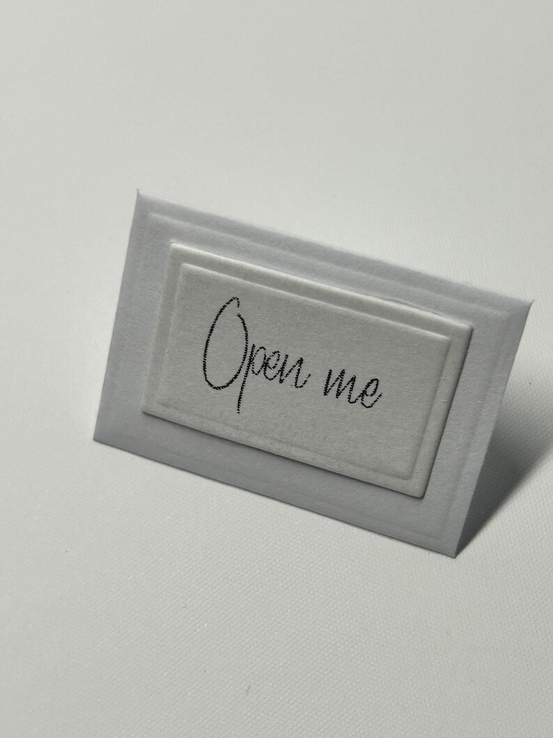 Puede incluir: Una tarjeta blanca con un borde en relieve y el texto "Open me" escrito en negro.