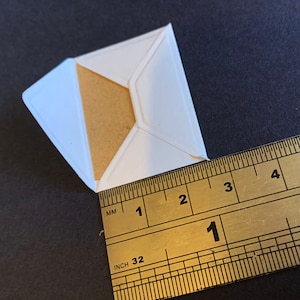 Puede incluir: Un sobre de papel blanco con un inserto de papel marr&oacute;n. El sobre se mide con una regla, mostrando 1 pulgada.