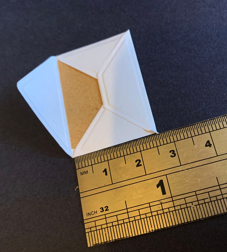Puede incluir: Un sobre de papel blanco con un inserto de papel marr&oacute;n. El sobre se mide con una regla, mostrando 1 pulgada.