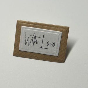 Puede incluir: Una peque&ntilde;a tarjeta marr&oacute;n con un rect&aacute;ngulo blanco en el centro. El rect&aacute;ngulo tiene las palabras "With Love" escritas en tinta negra.