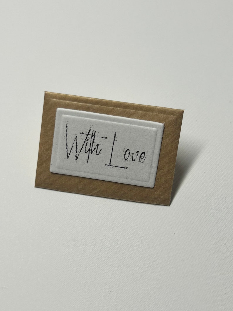 Puede incluir: Una peque&ntilde;a tarjeta marr&oacute;n con un rect&aacute;ngulo blanco en el centro. El rect&aacute;ngulo tiene las palabras "With Love" escritas en tinta negra.