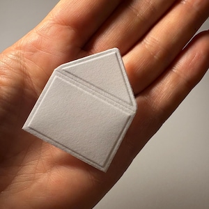 Puede incluir: Un sobre de papel blanco con una solapa doblada. El sobre es texturizado y tiene un dise&ntilde;o ligeramente en relieve.