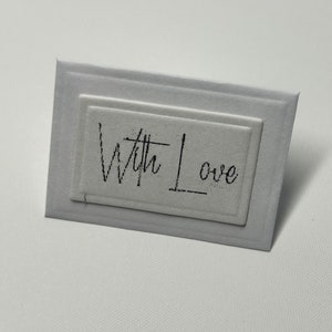 Puede incluir: Una tarjeta blanca con un borde negro y el texto "With Love" escrito en cursiva.