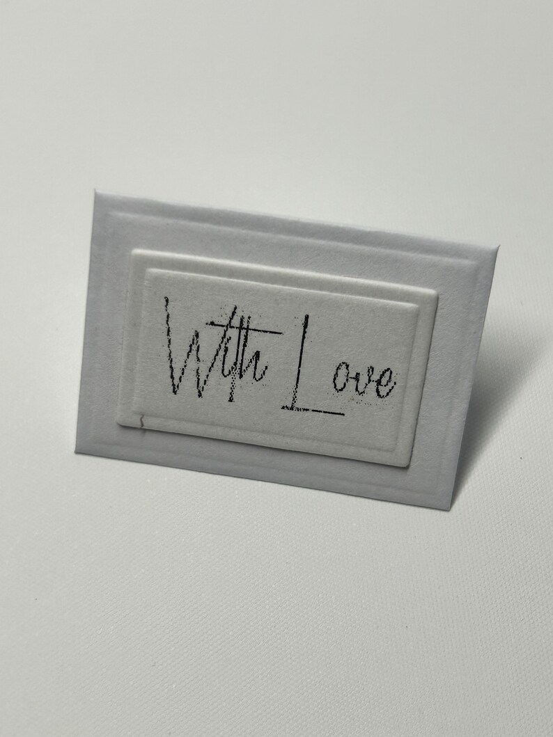 Puede incluir: Una tarjeta blanca con un borde negro y el texto "With Love" escrito en cursiva.