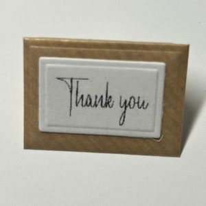 Puede incluir: Una peque&ntilde;a tarjeta marr&oacute;n con un rect&aacute;ngulo blanco en el centro. El rect&aacute;ngulo tiene las palabras "Gracias" escritas en cursiva negra.