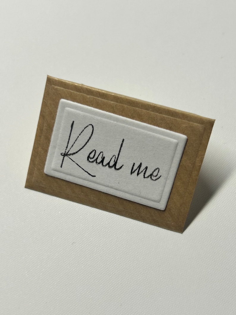 Puede incluir: Una peque&ntilde;a etiqueta de papel marr&oacute;n con un rect&aacute;ngulo blanco en el centro. El rect&aacute;ngulo tiene las palabras "Read me" escritas en cursiva negra.