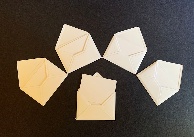 Puede incluir: Cinco sobres de papel blanco sobre un fondo negro. Los sobres est&aacute;n doblados en forma triangular y est&aacute;n dispuestos en forma de estrella.